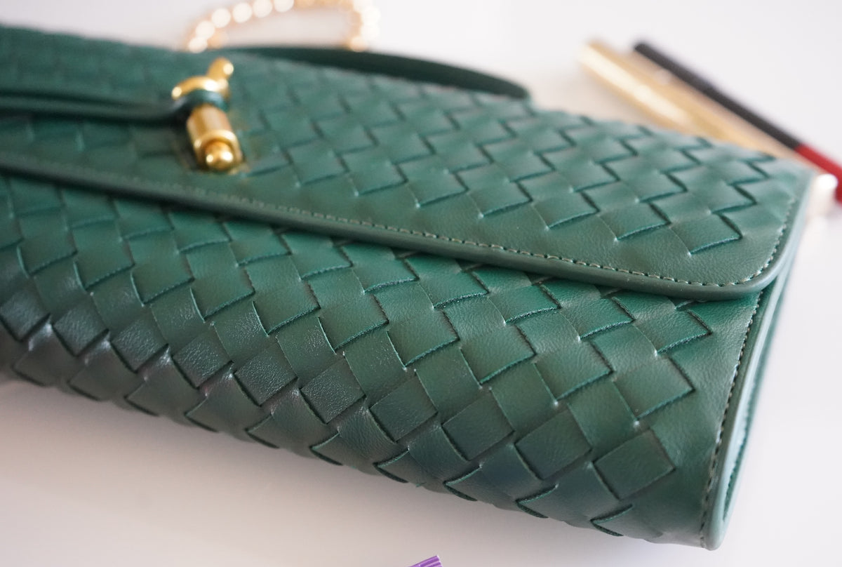 A close-up of a green woven leather clutch bag with gold-tone hardware, resting on a white surface with a blurred background.