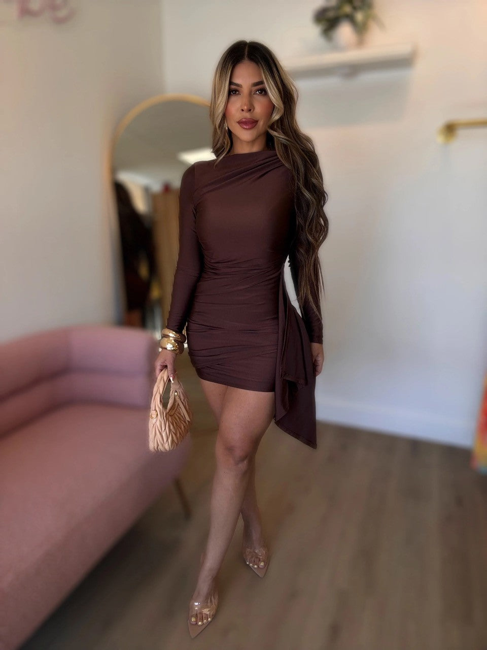 A woman models a fitted, long-sleeve mini dress in a deep chocolate brown color. The dress features an asymmetrical draped neckline and a draped side overlay.